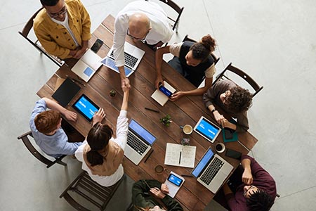 eight adults of various ages sitting a table with laptops, two of the people are shaking hands over the table